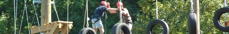 Zwei Kletternde mit Helm auf einem Element aus hängenden Autoreifen im Hochseilgarten