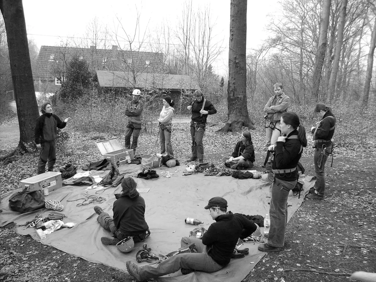 Schwarz-Weiß-Foto: Trainerinnen und Trainer in Klettergurten stehen und sitzen im Wald im Kreis um eine Plane mit Seilen, Helmen und Materialkisten
