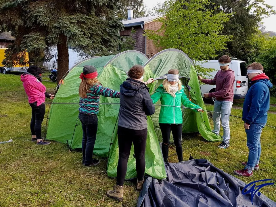 Teilnehmende mit Augenbinden bauen gemeinsam ein grünes Zelt auf einer Wiese auf