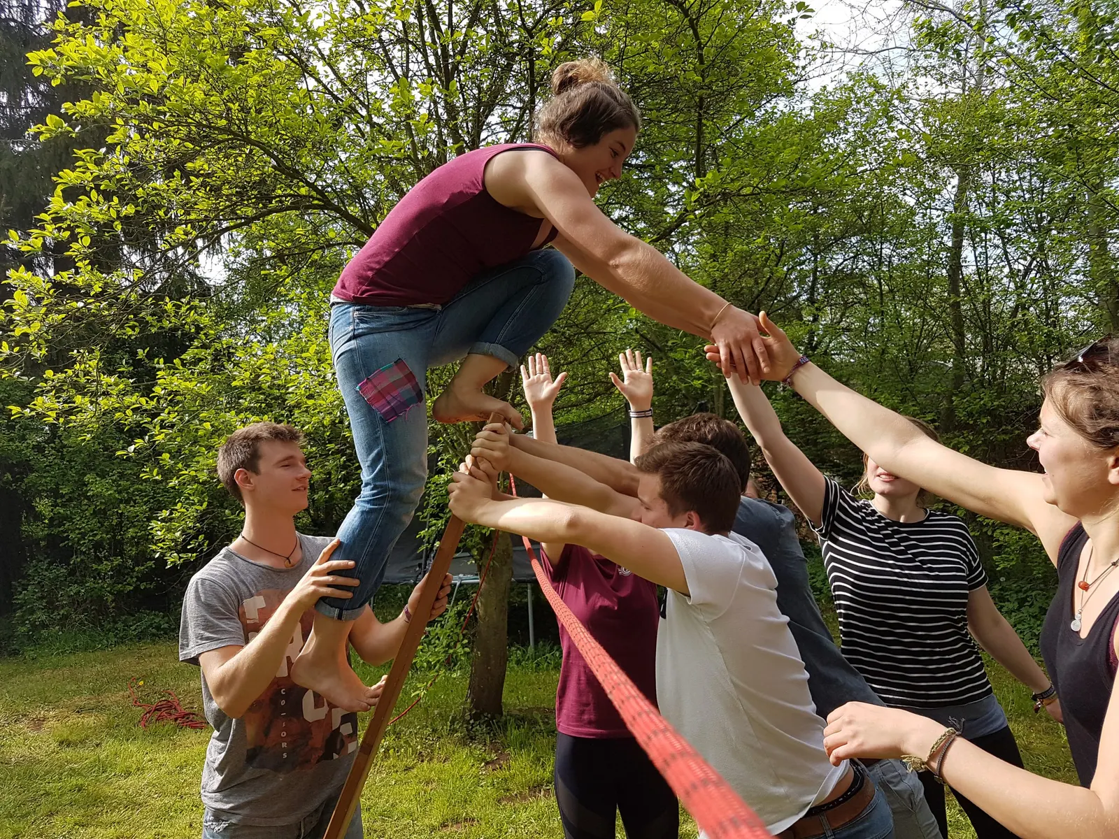 Teamaufgabe im Garten: Eine junge Frau steigt, von der Gruppe gestützt, auf einem Holzstab über ein gespanntes rotes Band und greift nach den ausgestreckten Händen auf der anderen Seite