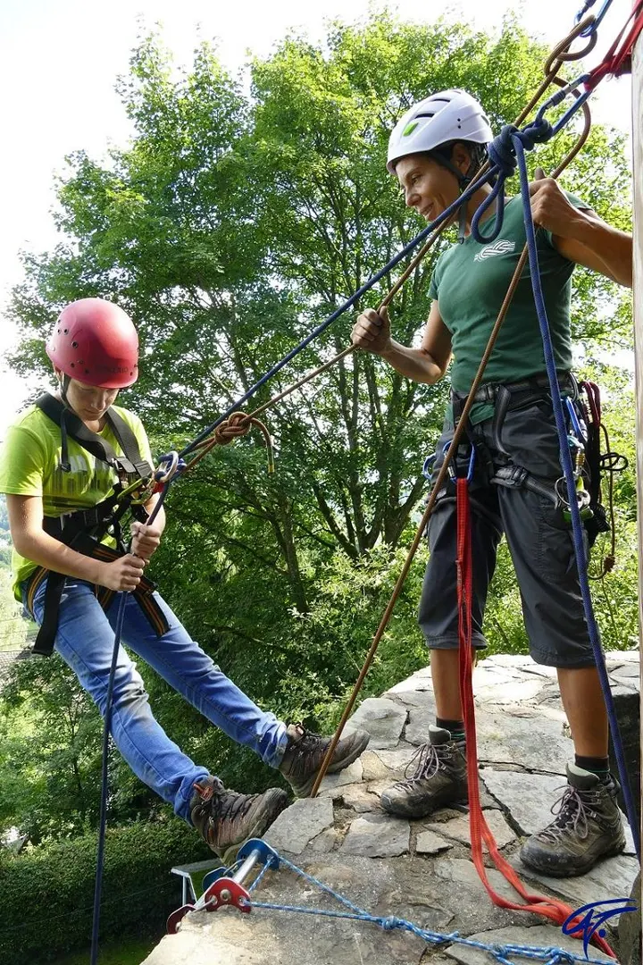 Eine Trainerin sichert ein Mädchen mit rotem Helm, das sich über eine Felskante abseilt