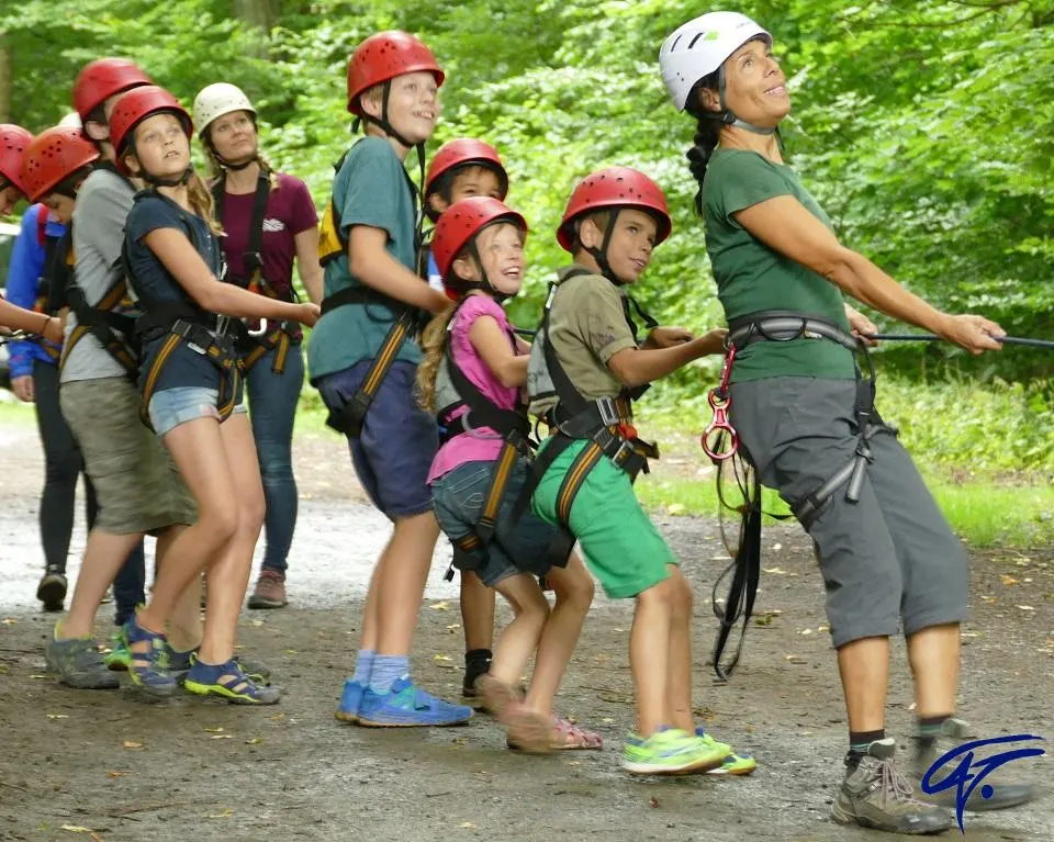 Kinder mit roten Kletterhelmen und Gurten ziehen gemeinsam mit einer Trainerin an einem Seil im Wald