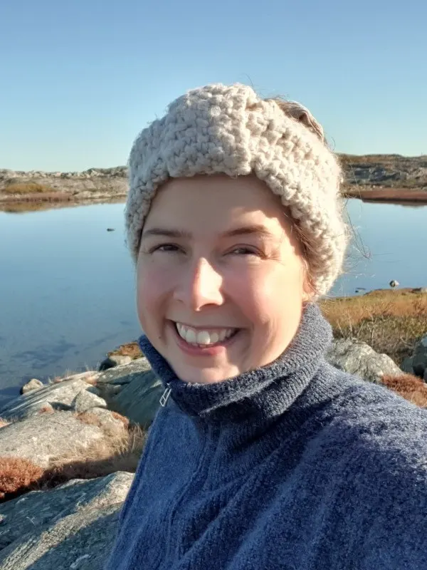 Tamar Sünder mit Strickstirnband und blauem Fleecepullover, lachend vor einem See in einer felsigen Landschaft
