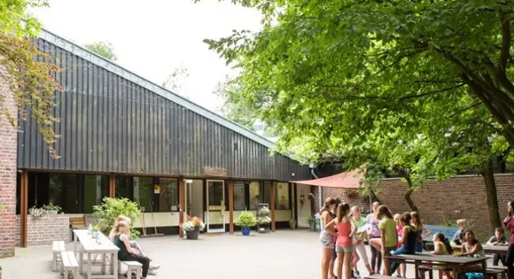 Innenhof der Jugendherberge Brüggen: Jugendliche stehen unter großen Laubbäumen vor einem dunklen Holzbau mit Sitzgruppen
