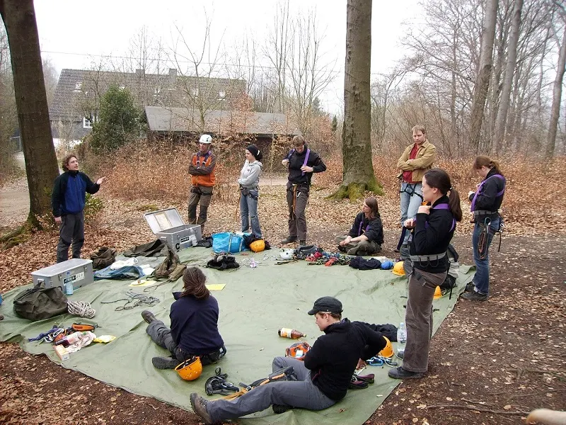 Eine Gruppe in Klettergurten steht und sitzt im winterlichen Wald rund um eine Plane mit Seilen, Helmen und Sicherungsmaterial und hört einer Person zu, die etwas erklärt