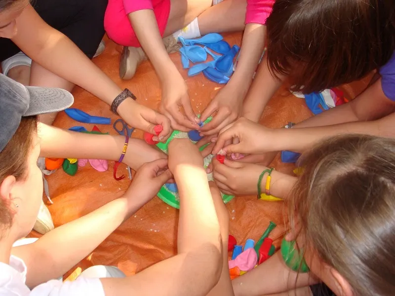 Viele Kinderhände greifen in der Mitte einer orangefarbenen Plane zusammen und knoten bunte Luftballons