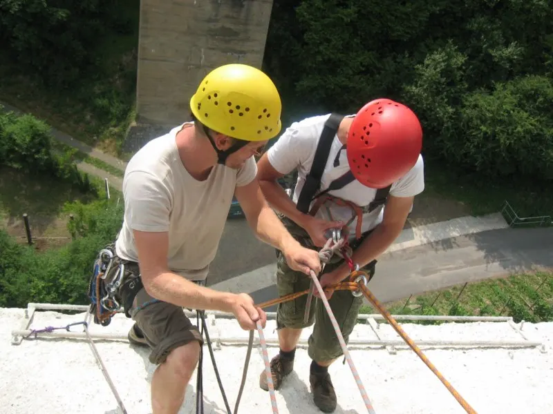 Ein Trainer mit gelbem Helm hilft einem Teilnehmer mit rotem Helm, sich an einem hohen Brückenpfeiler ins Abseilseil einzuhängen