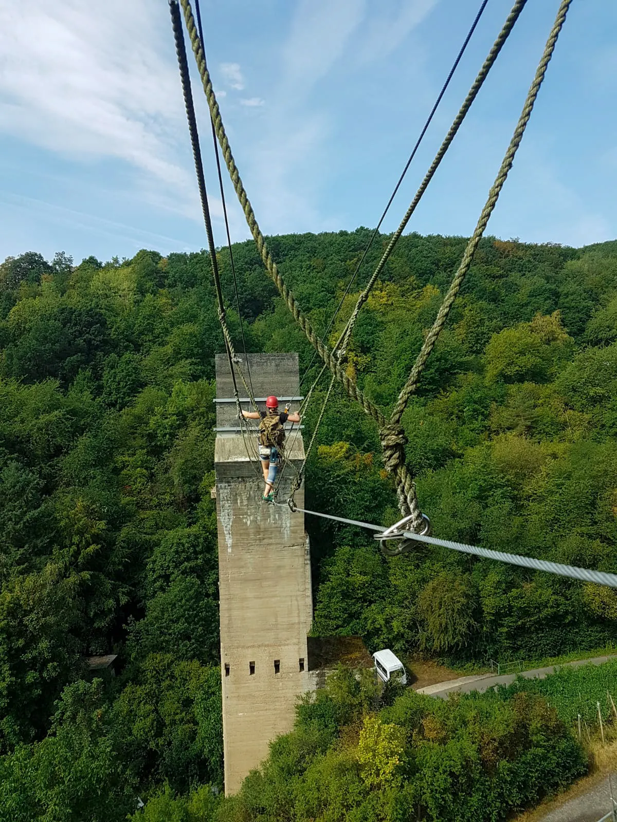 Eine Trainerin mit rotem Helm und Rucksack balanciert gesichert über ein Stahlseil auf den Betonpfeiler zu, tief unter ihr bewaldete Hänge bei Ahrweiler
