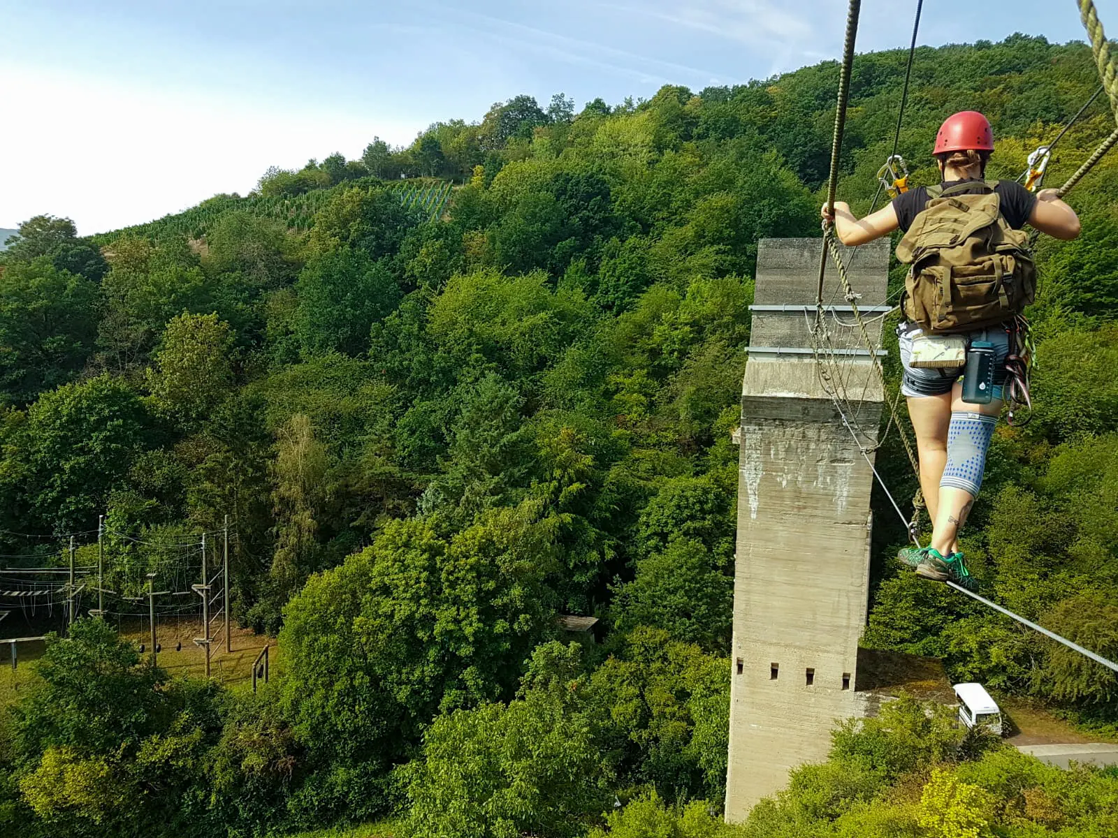 Eine angehende Hochseilgarten-Trainerin mit rotem Helm balanciert gesichert auf einem Stahlseil neben dem Kletterpfeiler, tief unter ihr das bewaldete Tal bei Ahrweiler