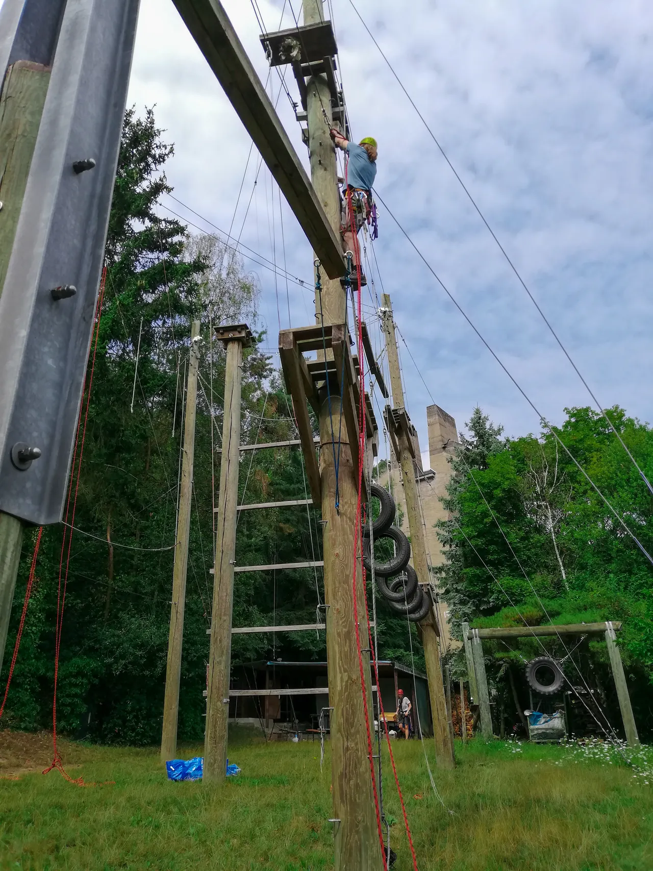 Jemand klettert mit Helm und Sicherungsseil einen Mast des Hochseilgartens hinauf, daneben eine Holzleiter und Autoreifen