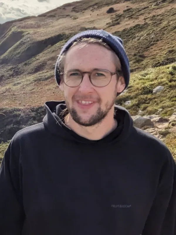Julian Arns mit Brille, Wollmütze und schwarzem Kapuzenpullover, lächelnd vor einer hügeligen Landschaft