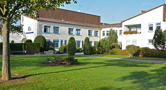 Jugendgästehaus Bad Neuenahr-Ahrweiler: weiße Gebäude mit blauen Fensterrahmen hinter einer sonnigen Rasenfläche mit Bäumen und Hecken