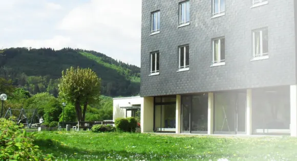 Jugendherberge Biedenkopf: schiefergedecktes Gebäude mit großer Glasfront, davor eine Blumenwiese, dahinter bewaldete Hügel