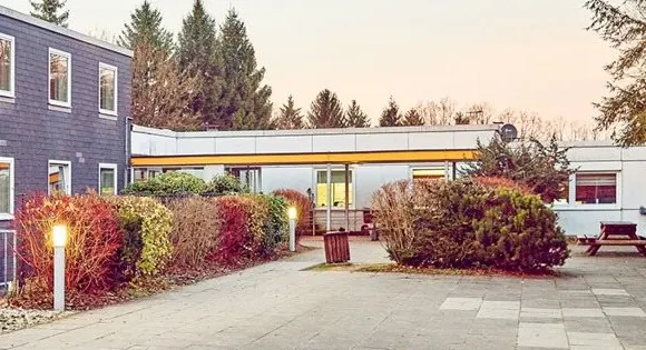 Jugendherberge Biggesee in der Abenddämmerung: Flachbau mit erleuchteten Fenstern, Wegleuchten und herbstlich gefärbten Sträuchern