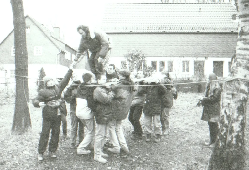 Schwarz-Weiß-Foto aus den Anfangsjahren: Eine Kindergruppe hilft einem Erwachsenen über ein zwischen Bäumen gespanntes Seil