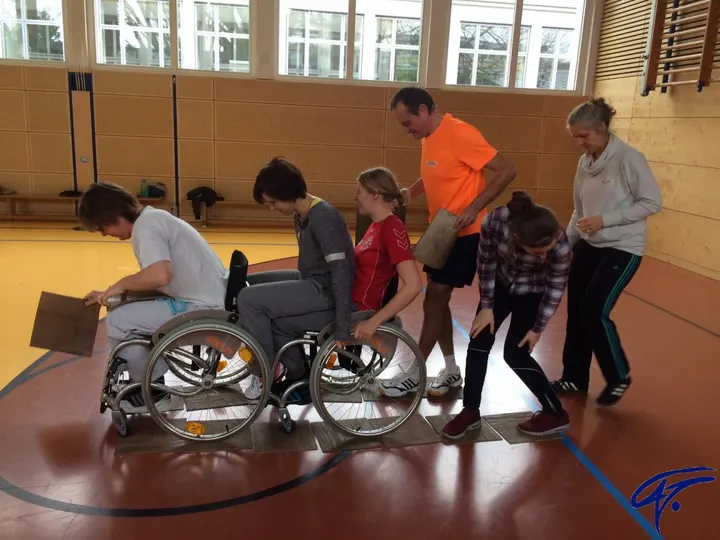 In einer Turnhalle fährt eine Person im Rollstuhl über Bretter, die die Gruppe vor ihr auslegt