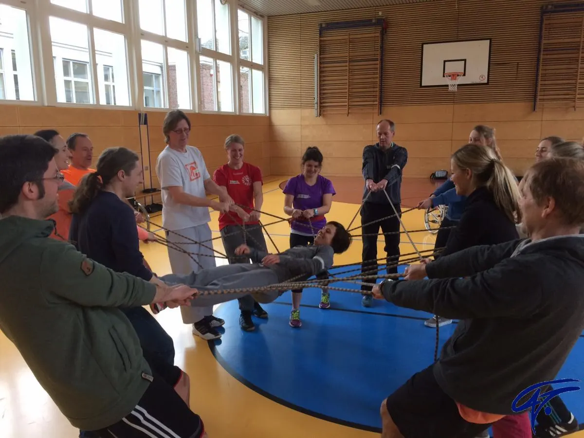 In einer Turnhalle spannen Erwachsene gemeinsam ein Netz aus Seilen, auf dem eine Person liegt
