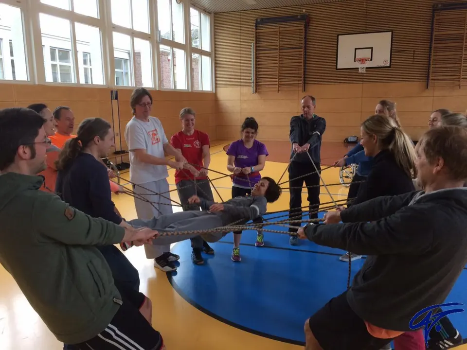 Erwachsene halten in einer Turnhalle gemeinsam ein Seilnetz, in dem eine Teilnehmerin liegt