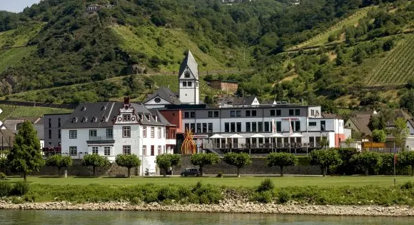 Jugendherberge Leutesdorf direkt am Rheinufer, dahinter ein Kirchturm und steile Weinberge