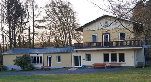 Schullandheim Norken: helles Haus mit Holzbalkon und Picknickbänken, umgeben von hohen, kahlen Bäumen