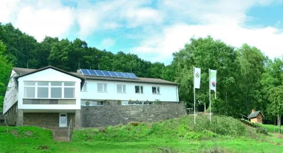 Schullandheim Kronenburg: weißes Haus mit Solaranlage und zwei Fahnen am grünen Hang vor dichtem Wald