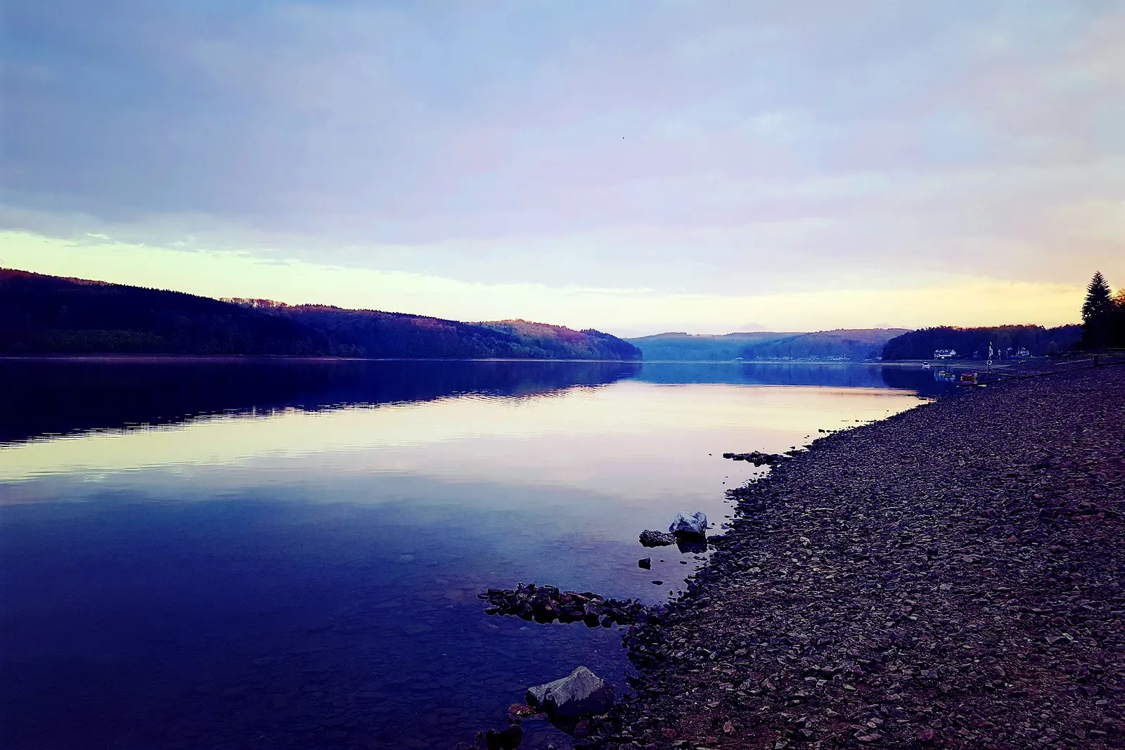 Abendstimmung am Sorpesee: Ein steiniges Ufer zieht sich am spiegelglatten Wasser entlang, dahinter bewaldete Hügel