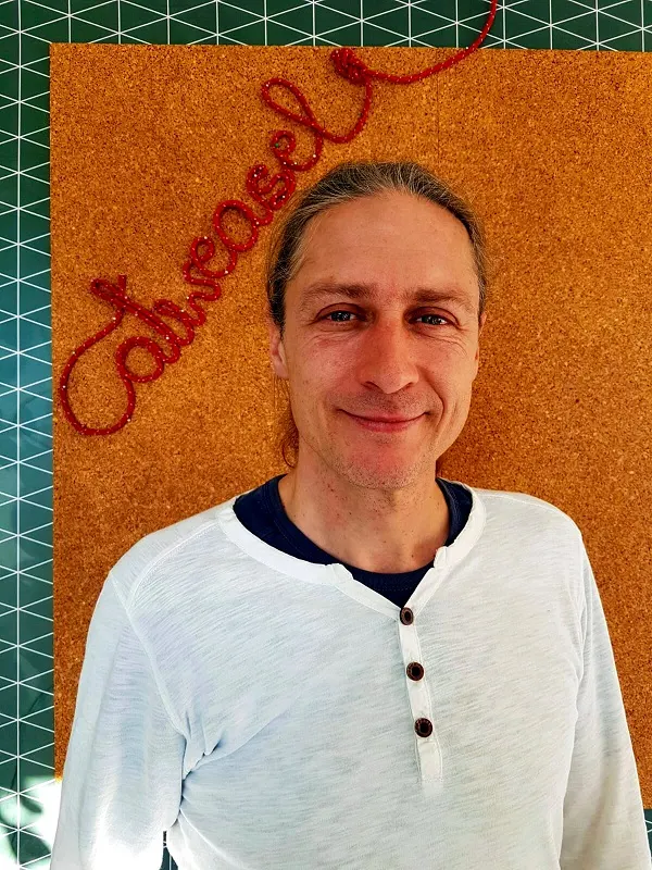 Stefan Gnirs, lächelnd, mit zurückgebundenen langen Haaren und weißem Shirt vor einer orangefarbenen Pinnwand mit dem Catweasel-Schriftzug aus Seil