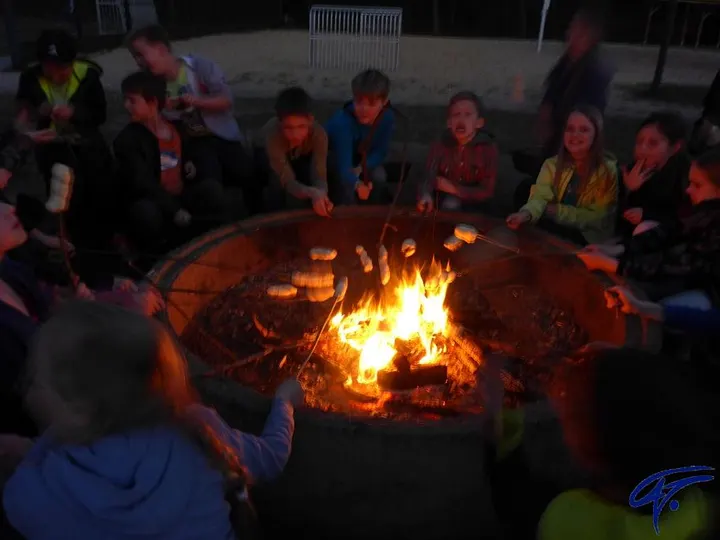 Kinder sitzen abends um eine Feuerschale und backen Stockbrot