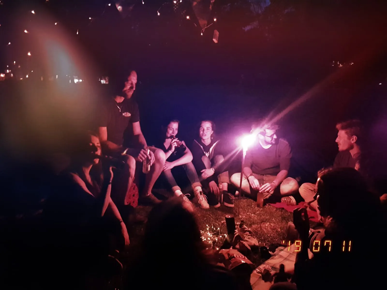TrainerInnen sitzen abends beim Sommerfest im Kreis auf der Wiese, im Hintergrund leuchten Lichterketten