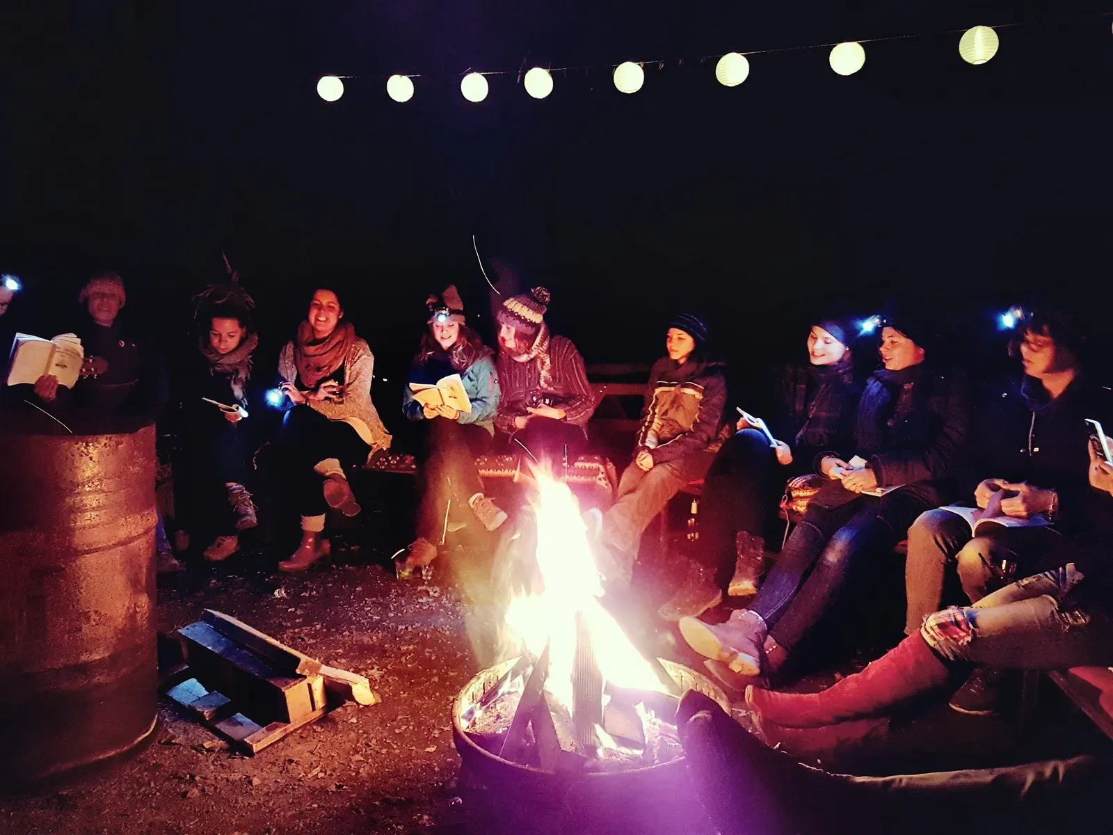 TrainerInnen singen nachts mit Stirnlampen und Liederheften am Lagerfeuer, über ihnen eine Lichterkette
