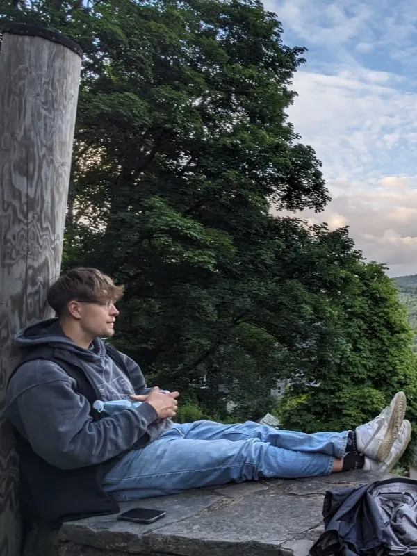 Yanick Schöne sitzt mit ausgestreckten Beinen auf einem Felsen unter hohen Bäumen und blickt in die Ferne