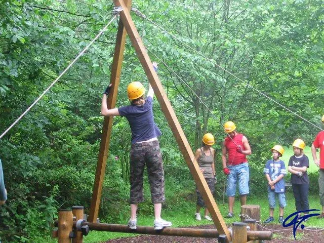 Eine Teilnehmerin mit Helm balanciert auf einer A-Brücke aus Holzbalken, die Gruppe hält die Seile