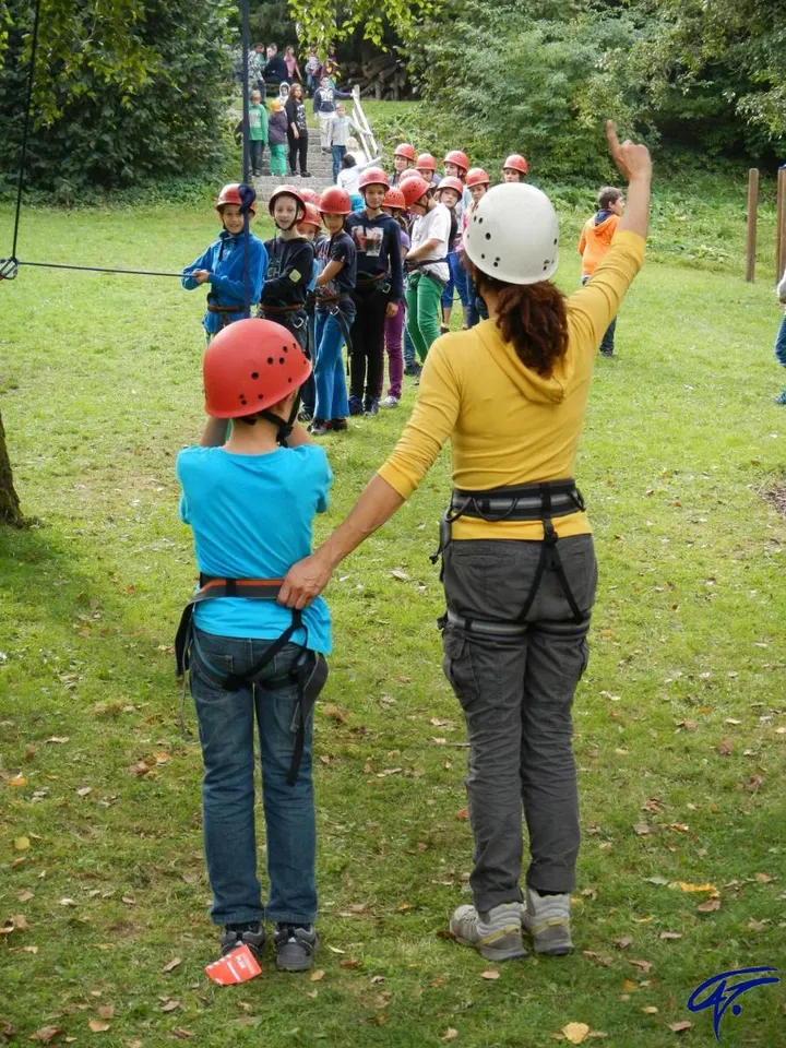 Kinder mit Helm und Gurt stehen in einer Reihe an der Hochziehstation, vorne eine Trainerin mit erhobenem Arm