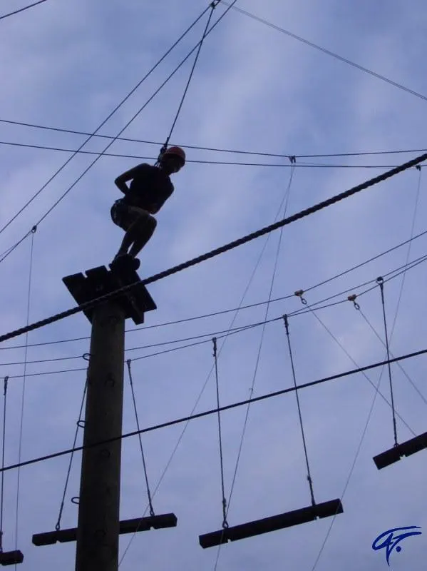 Eine Person steht im Gegenlicht auf der Spitze eines Hochseilgarten-Masts vor blauem Himmel