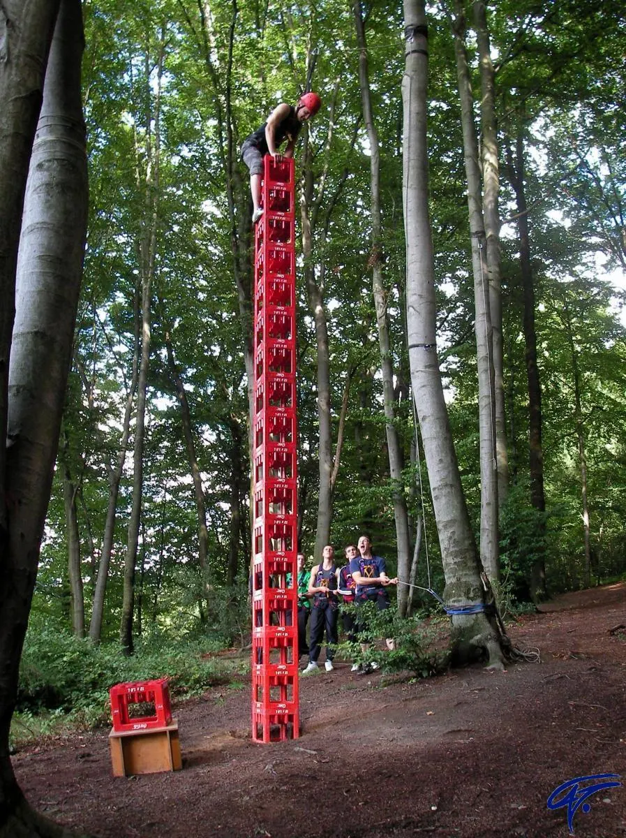 Ein Jugendlicher steht mit Helm auf einem hohen Turm aus roten Getränkekisten im Wald, gesichert von der Gruppe am Boden