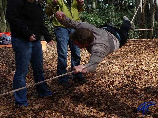 Ein Teilnehmer hängt im Herbstwald waagerecht an einer Niedrigseilkonstruktion, zwei Personen sichern ihn