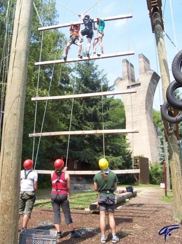 Ein Team steht oben auf der Riesenleiter, unten halten drei Jugendliche mit Helm die Sicherungsseile