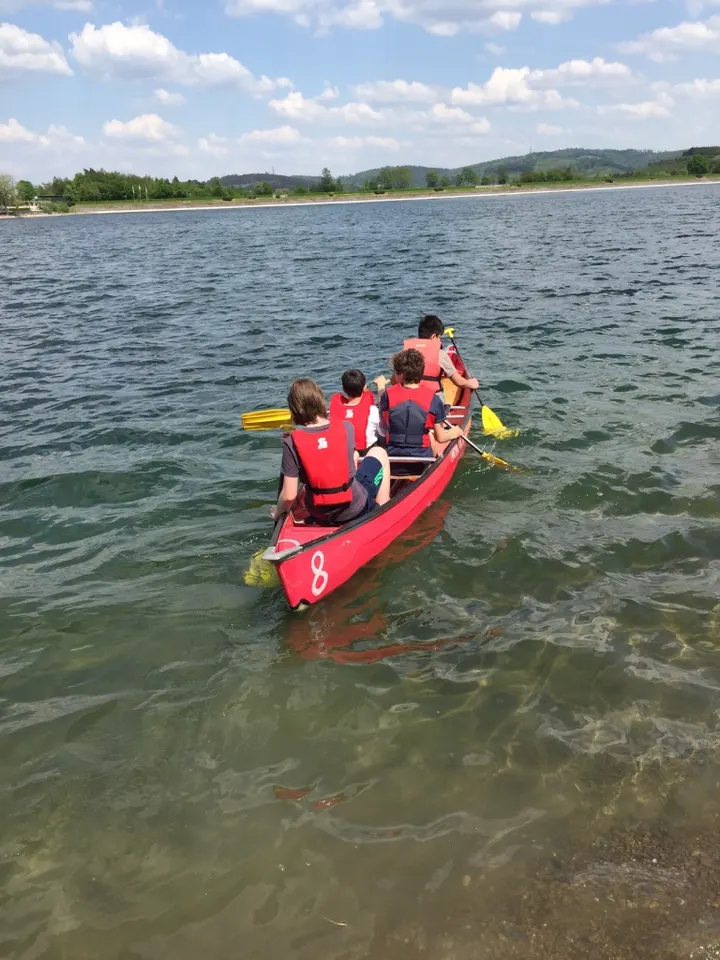 Drei Kinder mit roten Schwimmwesten paddeln in einem roten Kanu über den See