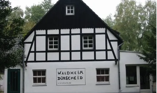 Das Waldheim Dürscheid: weißes Fachwerkhaus mit schwarzem Giebel und Namensschild an der Fassade, umgeben von Bäumen